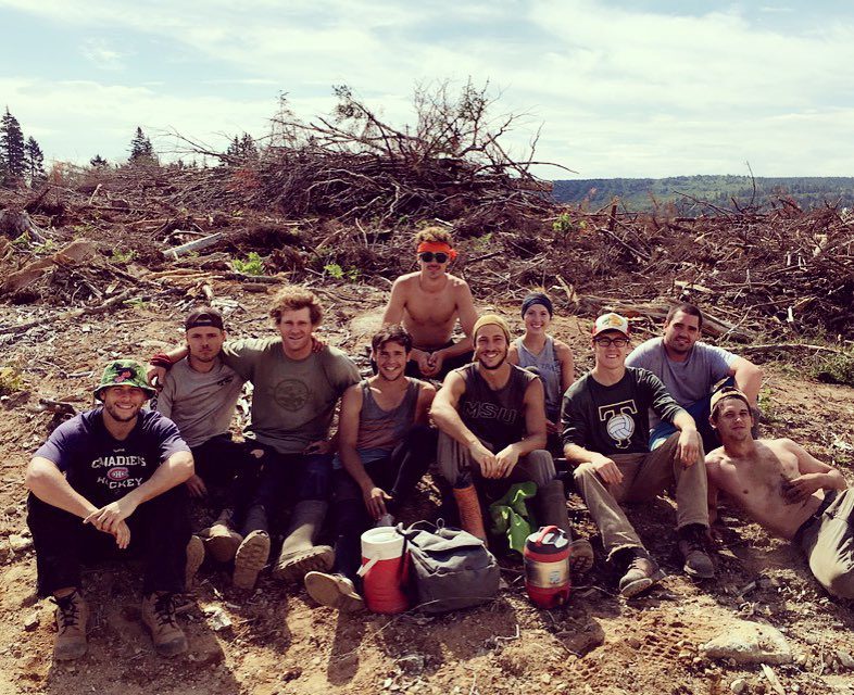 A tree-planting crew of twelve sitting on a cleared block on a sunny day, water jugs and boots in the foreground.